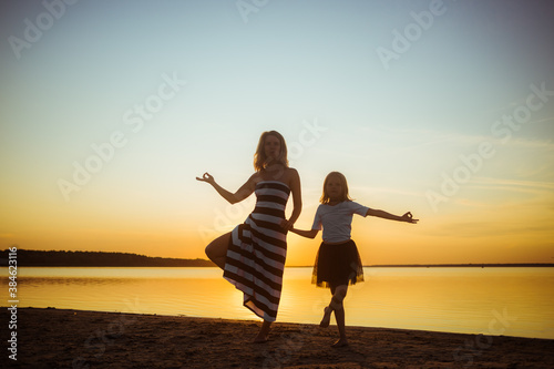 Mom with daughter in dresses are in a yoga pose on the lake at sunset. Leisure that strengthens the psychological health of the family and the body immunity through hardening.