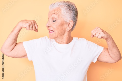 Senior beautiful woman with blue eyes and grey hair wearing classic white tshirt over yellow background showing arms muscles smiling proud. fitness concept.