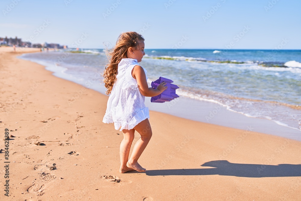Adorable blonde child on back view wearing summer dress. Holding beach bucket at the sand Adorable blonde child on back view wearing summer dress. Holding beach bucket at the sand