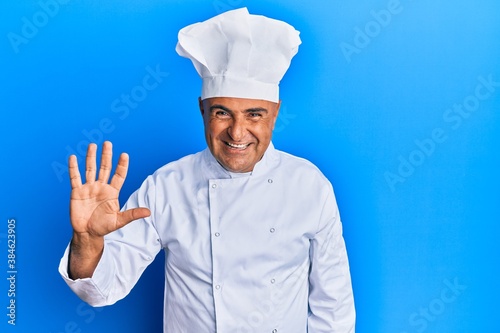 Mature middle east man wearing professional cook uniform and hat showing and pointing up with fingers number five while smiling confident and happy.