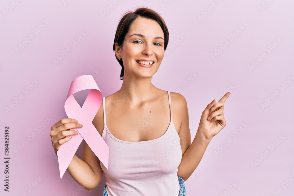 Young brunette woman with short hair holding pink cancer ribbon smiling happy pointing with hand and finger to the side Young brunette woman with short hair holding pink cancer ribbon smiling happy pointing with hand and finger to the side
