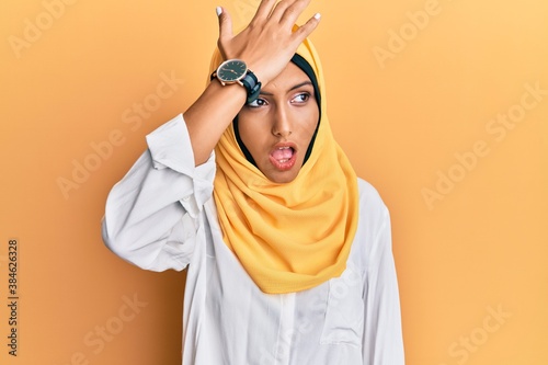 Young brunette arab woman wearing traditional islamic hijab scarf surprised with hand on head for mistake, remember error. forgot, bad memory concept.