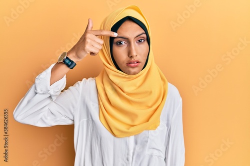 Young brunette arab woman wearing traditional islamic hijab scarf pointing unhappy to pimple on forehead, ugly infection of blackhead. acne and skin problem © Krakenimages.com
