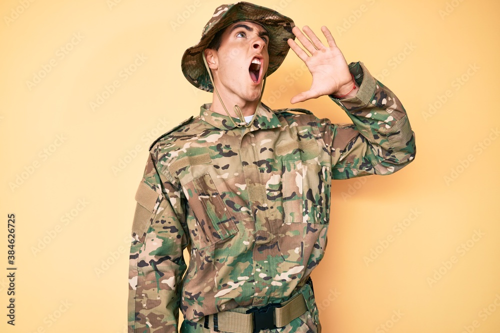 Young handsome man wearing camouflage army uniform shouting and screaming loud to side with hand on mouth. communication concept. Young handsome man wearing camouflage army uniform shouting and screaming loud to side with hand on mouth. communication concept.