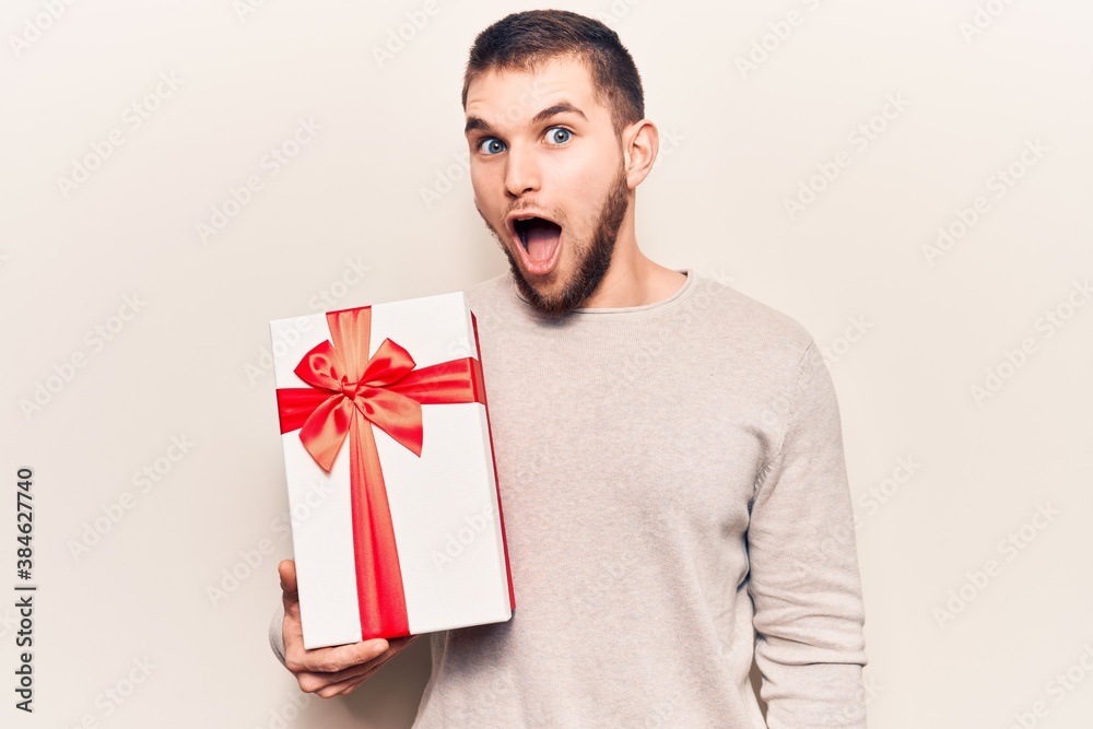 Young handsome man holding gift scared and amazed with open mouth for surprise, disbelief face Young handsome man holding gift scared and amazed with open mouth for surprise, disbelief face