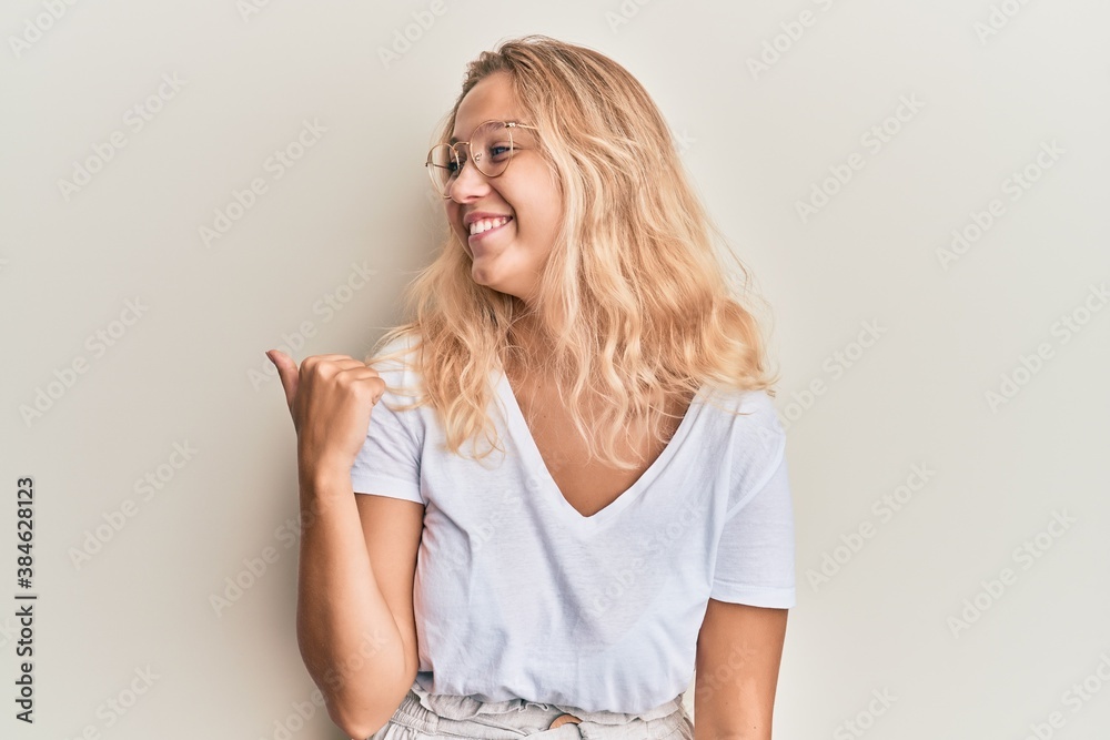Young blonde girl wearing casual clothes and glasses pointing thumb up to the side smiling happy with open mouth Young blonde girl wearing casual clothes and glasses pointing thumb up to the side smiling happy with open mouth