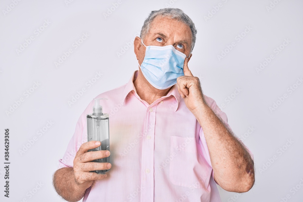Senior grey-haired man wearing medical mask holding hand sanitizer gel serious face thinking about question with hand on chin, thoughtful about confusing idea Senior grey-haired man wearing medical mask holding hand sanitizer gel serious face thinking about question with hand on chin, thoughtful about confusing idea