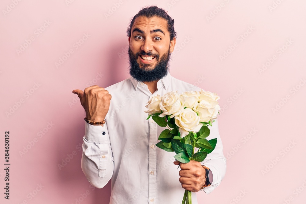 Young arab man holding flowers pointing thumb up to the side smiling happy with open mouth Young arab man holding flowers pointing thumb up to the side smiling happy with open mouth