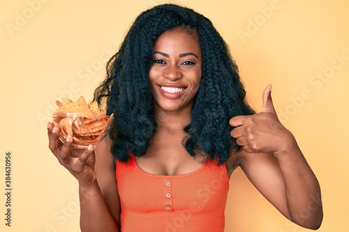 Beautiful african woman holding nachos potato chips smiling happy and positive  thumb up doing excellent and approval sign