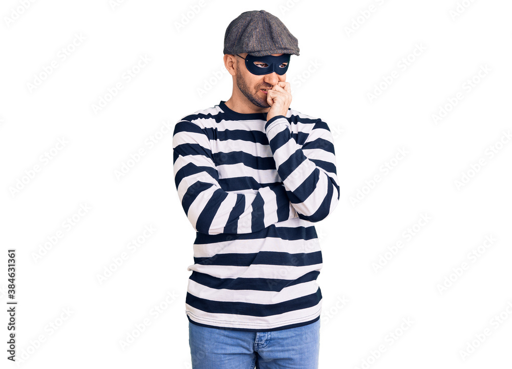 Young handsome man wearing burglar mask looking stressed and nervous with hands on mouth biting nails. anxiety problem. Young handsome man wearing burglar mask looking stressed and nervous with hands on mouth biting nails. anxiety problem.