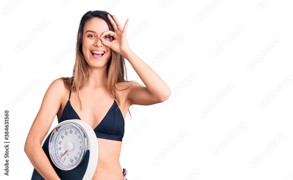Young beautiful girl wearing bikini holding weighing machine smiling happy doing ok sign with hand on eye looking through fingers Young beautiful girl wearing bikini holding weighing machine smiling happy doing ok sign with hand on eye looking through fingers