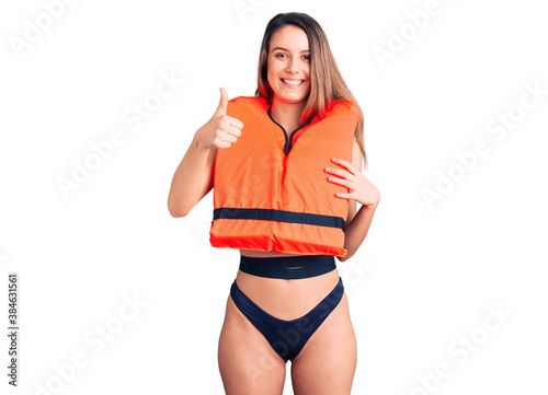 Young beautiful girl wearing life jacket smiling happy and positive, thumb up doing excellent and approval sign photo