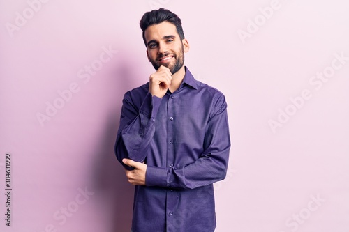 Young handsome man with beard wearing casual shirt looking confident at the camera smiling with crossed arms and hand raised on chin. thinking positive.