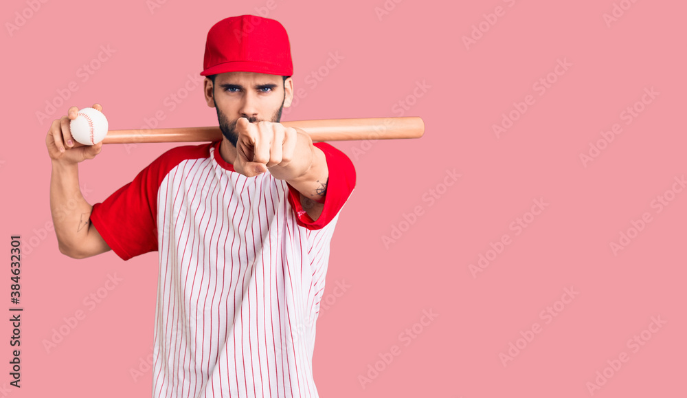 Young handsome man with beard playing baseball holding bat and ball pointing with finger to the camera and to you, confident gesture looking serious - obrazy, fototapety, plakaty 