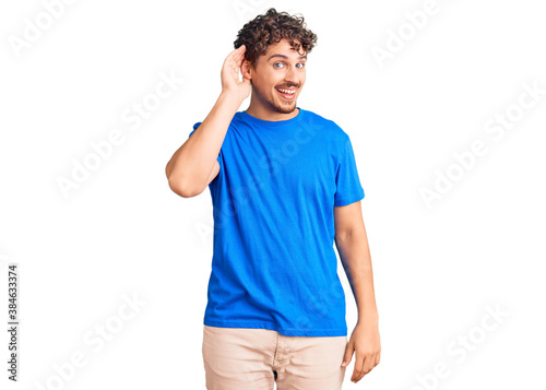 Young handsome man with curly hair wearing casual clothes smiling with hand over ear listening an hearing to rumor or gossip. deafness concept.