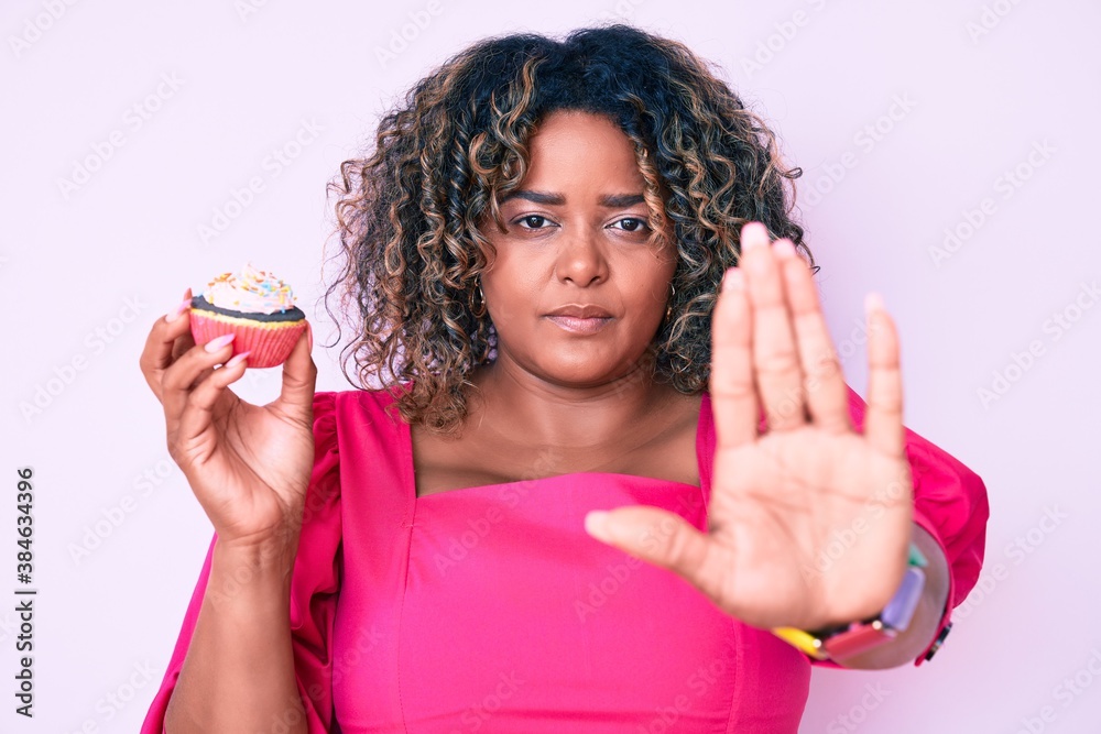 Young african american plus size woman holding cupcake with open hand doing stop sign with serious and confident expression, defense gesture Young african american plus size woman holding cupcake with open hand doing stop sign with serious and confident expression, defense gesture