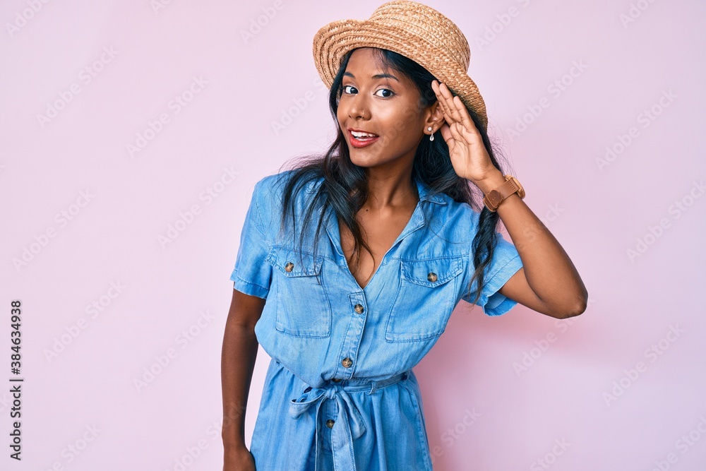 Young indian girl wearing summer hat smiling with hand over ear listening and hearing to rumor or gossip. deafness concept. Young indian girl wearing summer hat smiling with hand over ear listening and hearing to rumor or gossip. deafness concept.