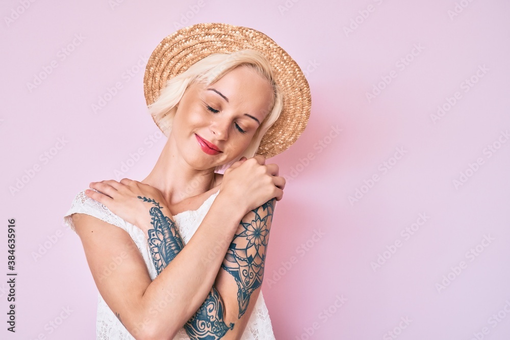 Young blonde woman with tattoo wearing summer hat hugging oneself happy and positive, smiling confident. self love and self care Young blonde woman with tattoo wearing summer hat hugging oneself happy and positive, smiling confident. self love and self care