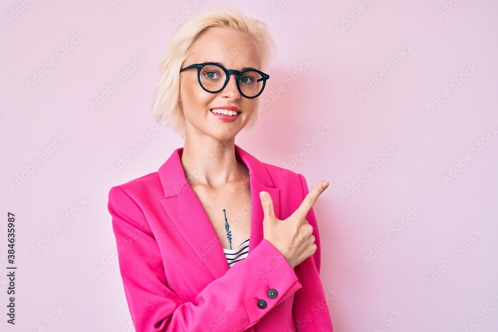 Young blonde woman with tattoo wearing business clothes and glasses smiling cheerful pointing with hand and finger up to the side Young blonde woman with tattoo wearing business clothes and glasses smiling cheerful pointing with hand and finger up to the side