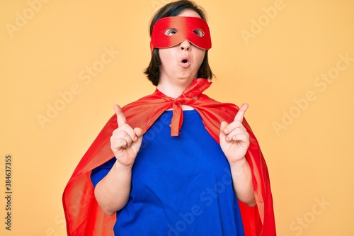 Brunette woman with down syndrome wearing super hero costume amazed and surprised looking up and pointing with fingers and raised arms.