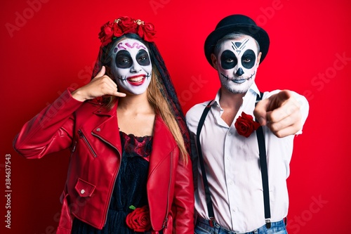 Couple wearing day of the dead costume over red smiling doing talking on the telephone gesture and pointing to you. call me.