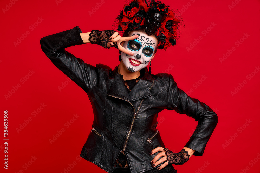 Dead gir with sugar skull makeup with a wreath of flowers on her head and skull, wearth lace gloves and showing victory sign isolated on red background.Concept of Halloween or La Calavera Catrina Dead gir with sugar skull makeup with a wreath of flowers on her head and skull, wearth lace gloves and showing victory sign isolated on red background.Concept of Halloween or La Calavera Catrina