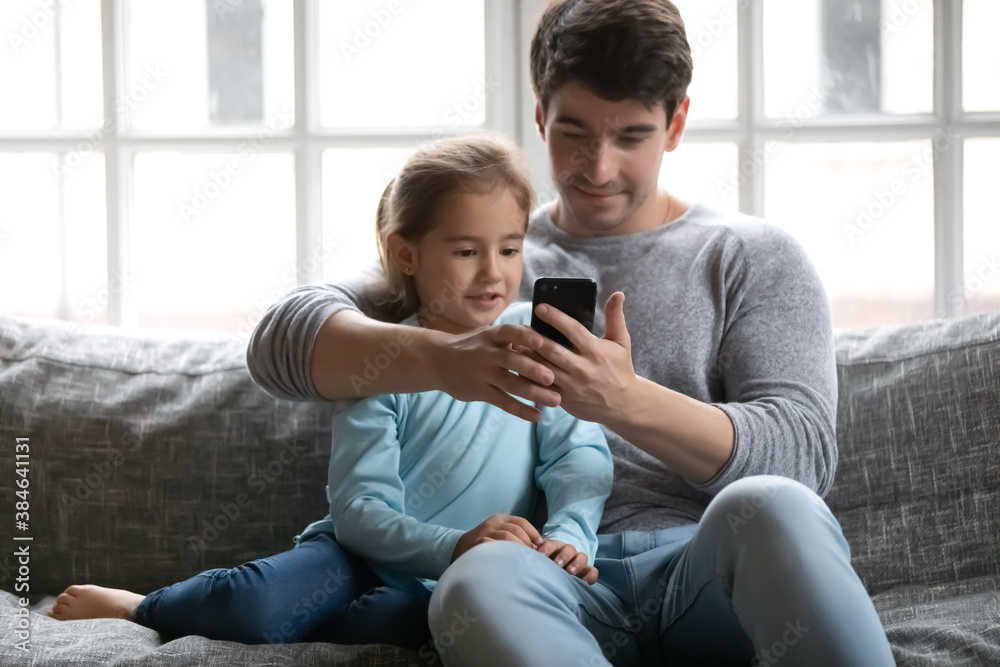 Caring young Caucasian father and small daughter sit relax on sofa in living room use smartphone together. Loving dad and little preschooler girl child rest on couch at home watch video on cellphone. Caring young Caucasian father and small daughter sit relax on sofa in living room use smartphone together. Loving dad and little preschooler girl child rest on couch at home watch video on cellphone.