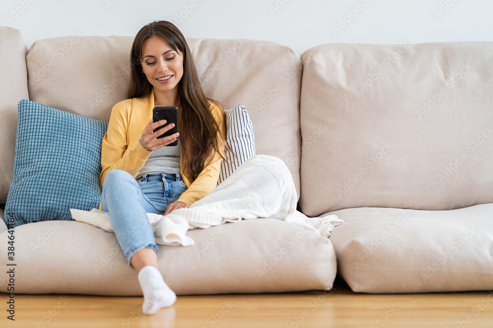 Smiling millennial woman relax sit on couch at home, using phone, texting chatting with friend. Happy girl resting on sofa hold smartphone, shopping online, check mobile application or social media. Smiling millennial woman relax sit on couch at home, using phone, texting chatting with friend. Happy girl resting on sofa hold smartphone, shopping online, check mobile application or social media.