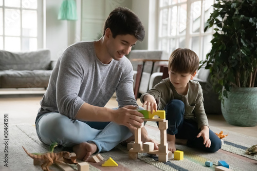 Loving young Caucasian father sit on floor at home play build with bricks with little son. Happy caring dad and small preschooler boy child have fun engaged in game activity in living room together.