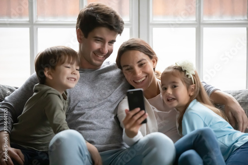 Happy young parents sit relax on couch at home watch funny video on cellphone with two small kids. Overjoyed loving family with little children rest enjoy weekend use smartphone gadget together.