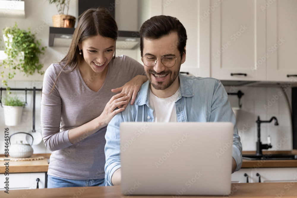 Happy young loving family couple looking at laptop screen, reading e-mail with good news. Smiling handsome man showing funny video on computer to pretty joyful wife, sitting at table in kitchen. Happy young loving family couple looking at laptop screen, reading e-mail with good news. Smiling handsome man showing funny video on computer to pretty joyful wife, sitting at table in kitchen.