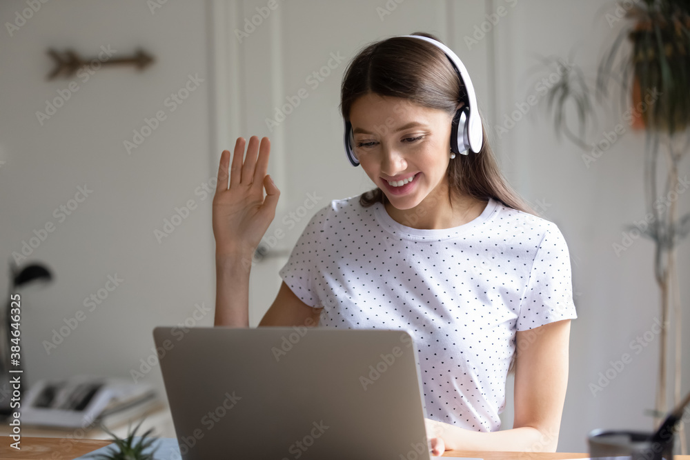 Smiling pretty young caucasian woman in modern earphones looking at laptop screen, making hello gesture, waving hand greeting colleagues at online video call or virtual event at home office. Smiling pretty young caucasian woman in modern earphones looking at laptop screen, making hello gesture, waving hand greeting colleagues at online video call or virtual event at home office.
