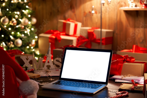 Laptop computer with white blank empty mock up screen on Merry Christmas table with presents gifts boxes, decorated Xmas tree in cozy Santa house background. Ecommerce website online shopping delivery
