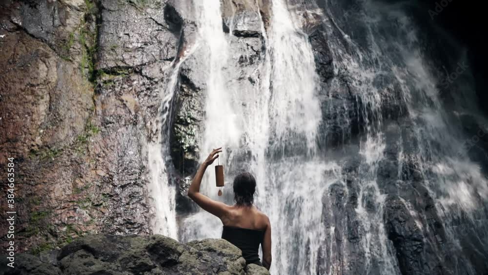 Young caucasian woman meditates while sitting on a rock in front of a waterfall with wooden wind chimes in her hands in a summer morning Young caucasian woman meditates while sitting on a rock in front of a waterfall with wooden wind chimes in her hands in a summer morning