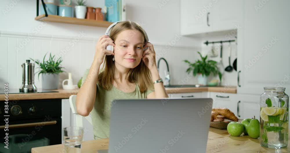Young Woman Puts on Headphones and Talks Online by Video Call using Laptop in Kitchen at Breakfast in Morning. Study or Remote Home Office Job at COVID-19 Coronavirus Pandemic. 4K Medium Shot Young Woman Puts on Headphones and Talks Online by Video Call using Laptop in Kitchen at Breakfast in Morning. Study or Remote Home Office Job at COVID-19 Coronavirus Pandemic. 4K Medium Shot