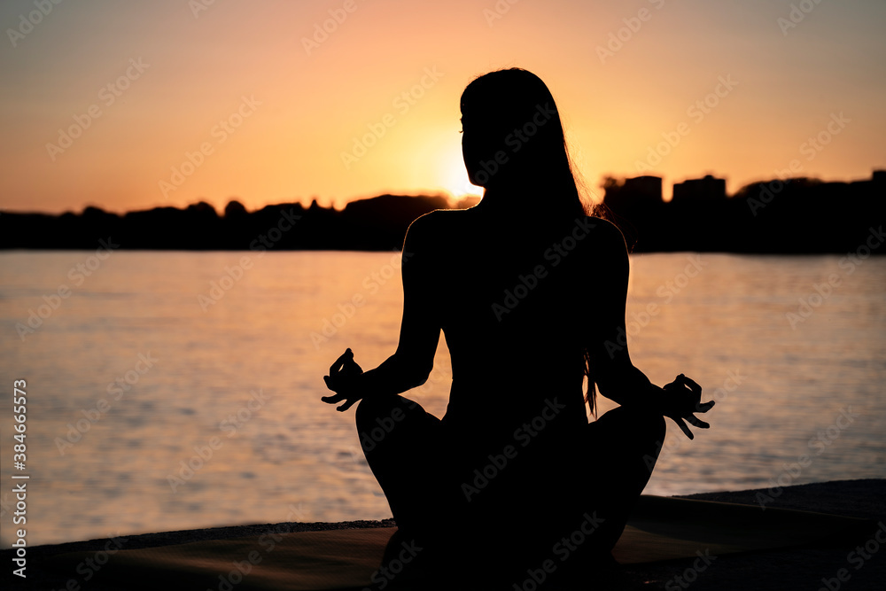 Silhouette of young woman meditating by the river at sunset. Peace and tranquil.