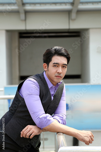 Southeast Asian young office business man wearing vest lean on railing stand look at camera confident expression outdoor premises