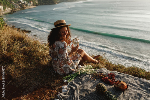 Young curly hair woman in straw hat sit on the edge of cliff with ocean view and drink water from stylish eco botle, decorated with macrame, enjoy picnic near the sea