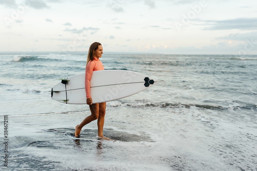 Young attractive surfer woman with white board at sunset on the ocean. Bali Indonesia. Summer time, sports, travel content.