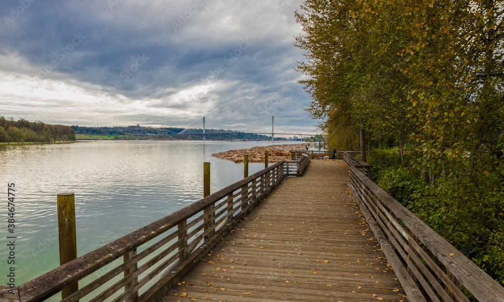 A wooden promenade along the Fraser River in Coquitlam City, yellowed trees along the bank, a bridge across the river on the horizon