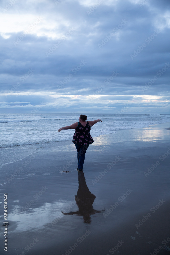 Woman pirouettes gracefully on the beach after sunset. Light still showing from between clouds. Reflection on sand.