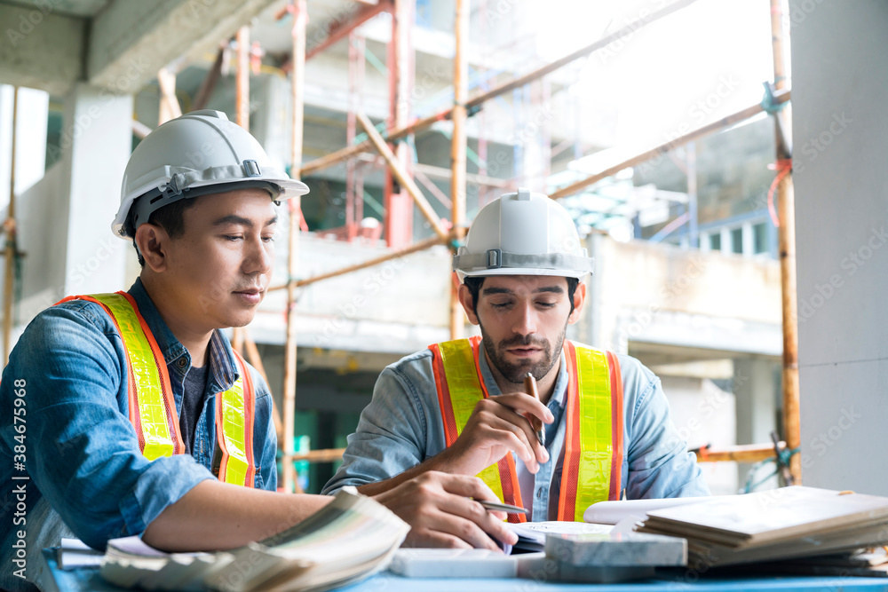 Young Asian engineers and caucasian engineer colleagues at site inspector and discuss together using notebook computer and paper blueprint at building construction site.home renovation ideas concept Young Asian engineers and caucasian engineer colleagues at site inspector and discuss together using notebook computer and paper blueprint at building construction site.home renovation ideas concept