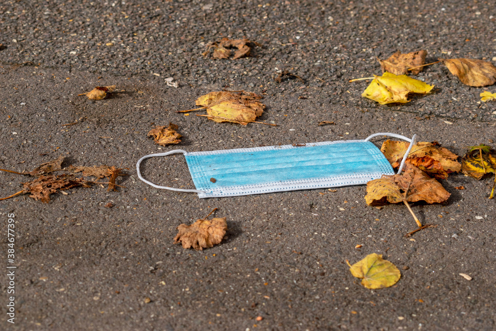 Obraz premium Blue disposable surgical mask on the cement floor in the middle of yellow leaves showing a new type of pollution during the pandemic we are living (Covid-19).