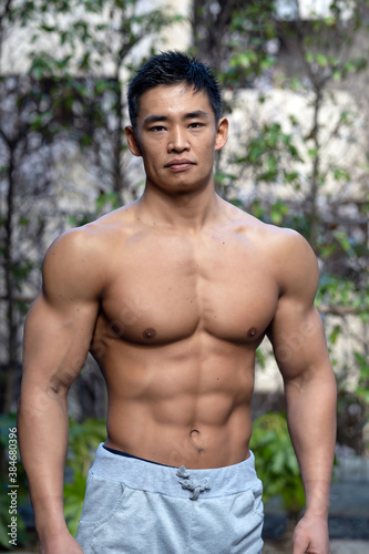 A young Asian bodybuilder poses shirtless outdoors showing off a muscular torso