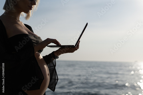silhouette of a pregnant woman on the background of the ocean, is typing text on a laptop.