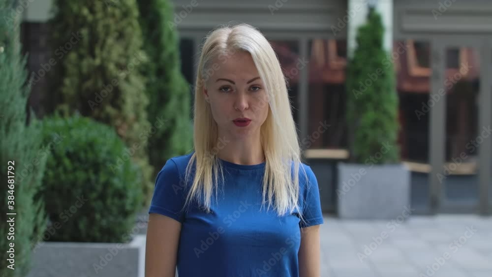 Portrait of a charming young woman looking at the camera and talking to someone. Blonde in a blue blouse outdoors on the background of the street. Slow motion. Close up. Portrait of a charming young woman looking at the camera and talking to someone. Blonde in a blue blouse outdoors on the background of the street. Slow motion. Close up.
