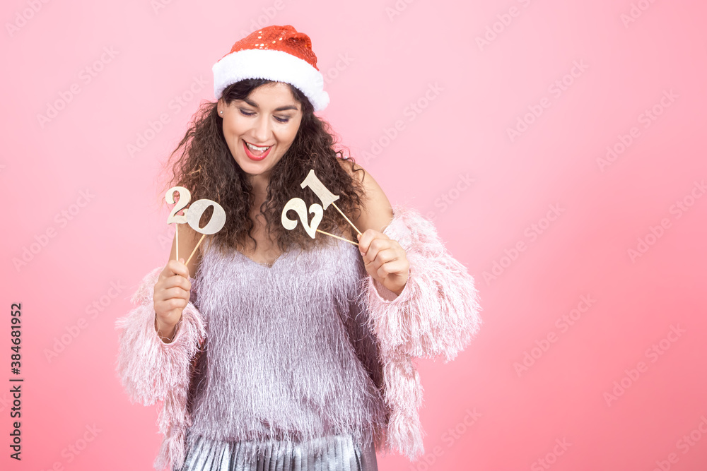 Young woman in a Christmas cap on a pink background holding a wooden number 2021 in her hand Young woman in a Christmas cap on a pink background holding a wooden number 2021 in her hand