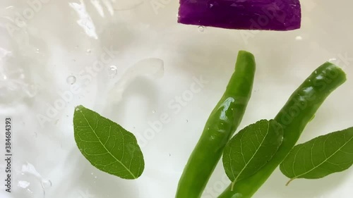 Pouring water and dropping chopped eggplant vegetable and onions slices into a bowl with leaves and green chilies creating beautiful splashes bubbles and ripples 