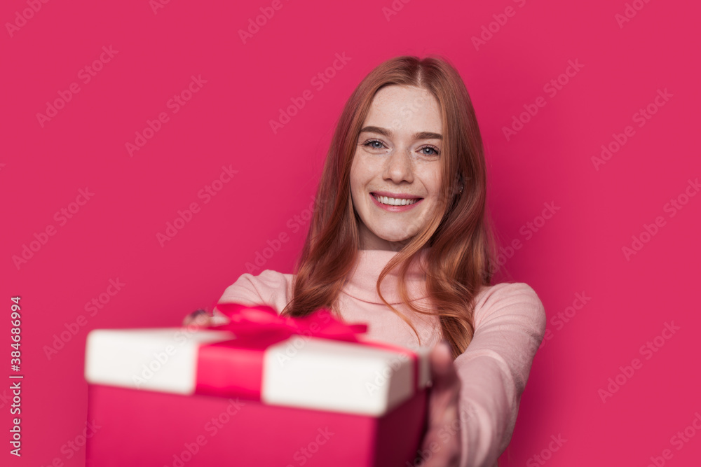 Charming ginger woman with freckles is giving a present at the camera smiling on a pink wall at studio Charming ginger woman with freckles is giving a present at the camera smiling on a pink wall at studio