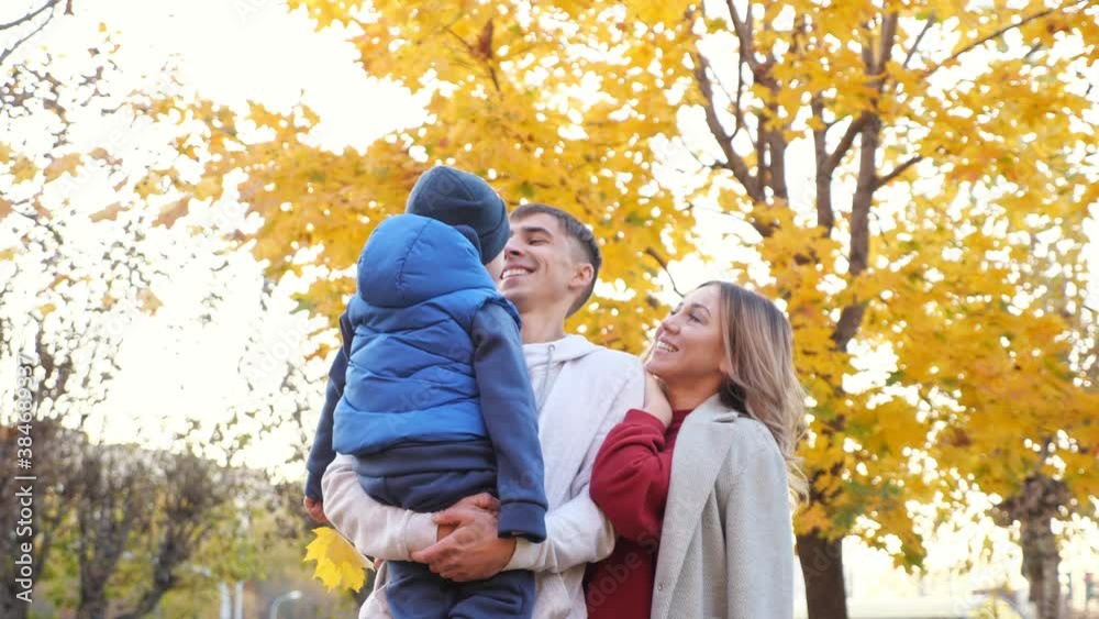 Cheerful family of mommy daddy and cute little son hug posing for camera in picturesque autumn city garden on nice warm day low angle shot Cheerful family of mommy daddy and cute little son hug posing for camera in picturesque autumn city garden on nice warm day low angle shot